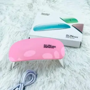 Mini lámpara UV portátil para uñas en color rosado brillante con cable USB sobre superficie peluda blanca y caja del producto de fondo.